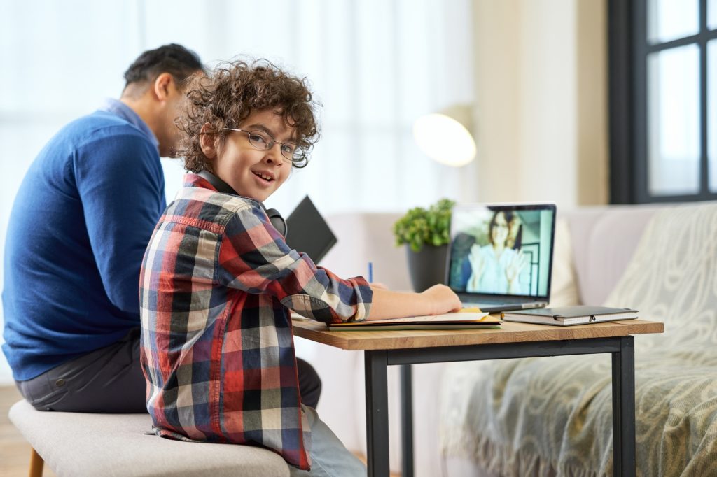 Study at your place. Cheerful latin boy wearing glasses sitting at the desk together with his father